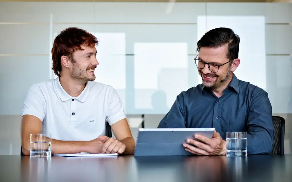 MID Mentoring | Zwei Personen im Gespräch am Tisch mit Tablet und Wassergläsern | MID GmbH