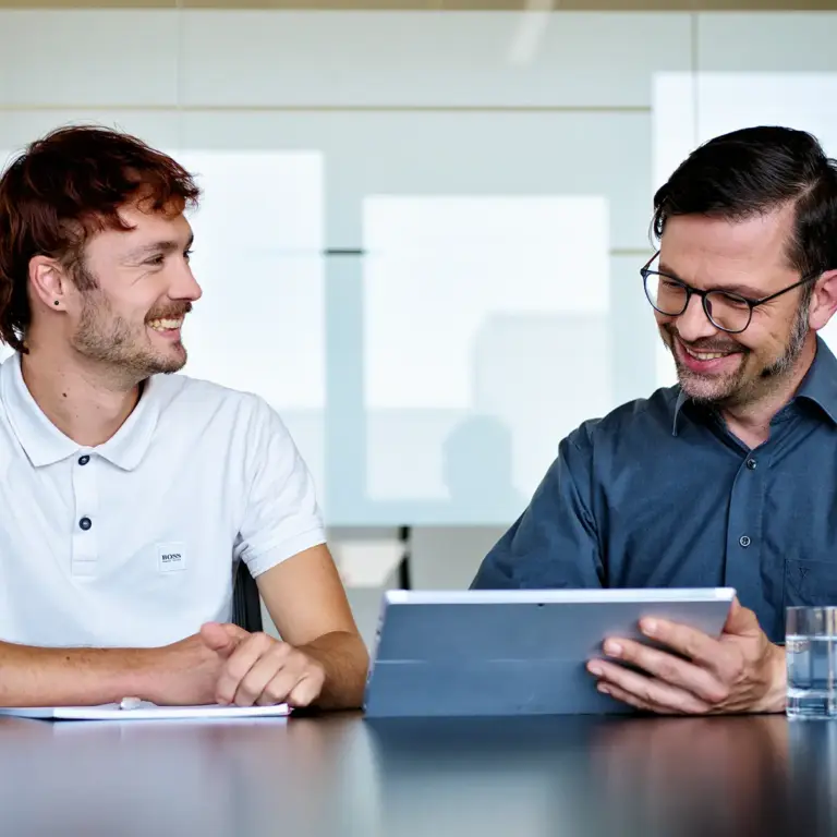 MID Mentoring | Zwei Personen im Gespräch am Tisch mit Tablet und Wassergläsern | MID GmbH