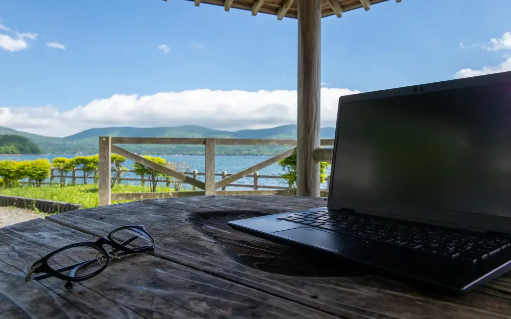 MID Mentoring | Laptop und Brille auf einem Holztisch unter einem Pavillon mit Blick auf einen See, Berge und blauen Himmel | MID GmbH