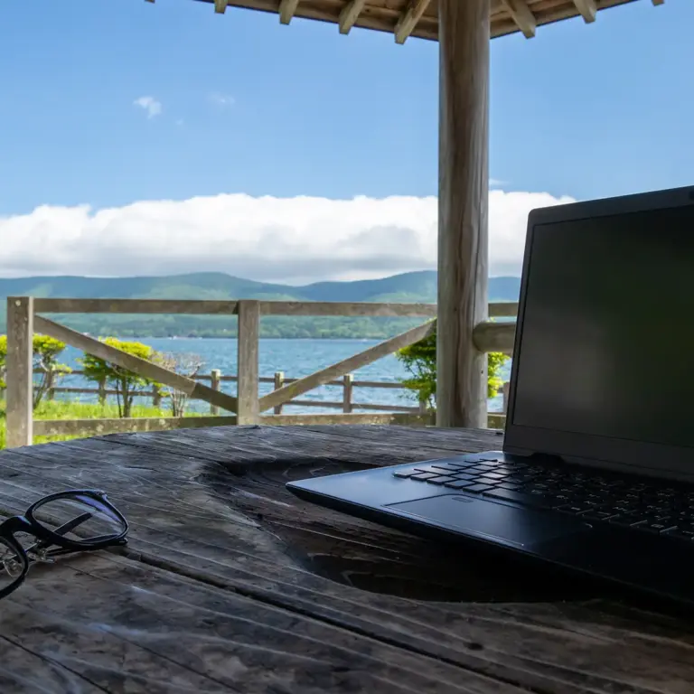MID Mentoring | Laptop und Brille auf einem Holztisch unter einem Pavillon mit Blick auf einen See, Berge und blauen Himmel | MID GmbH